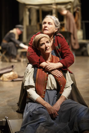 Mother Courage in red coat and looking off to the left behind Kattrin has her arms around the daughter's shoulder, Kattrin in Bohemian peasant dress and red vest looks off to the right, her hands dangling in her lap